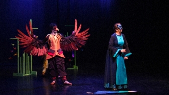 Hombre disfrazado de pájaro a la izquierda con las alas abiertas observando a una mujer con máscara de gata.