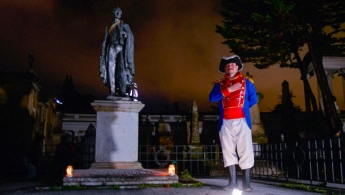 Hombre con disfraz de la campaña libertadora evocando el saludo a la patria a su lado derecho es encuentra la estatua de un prócer y se encuentran en el cementerio central de noche.
