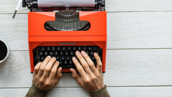 Fotografía en toma cenital de una maquina de escribir de color rojo en la cual hay unas manos adultas dispuestas a escribir y al lado una taza de tinto, todo sobre una mesa de madera blanca.