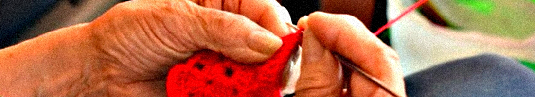 Fotografía en primer plano de las manos de una señora de la tercera edad tejiendo con lana de color rojo.