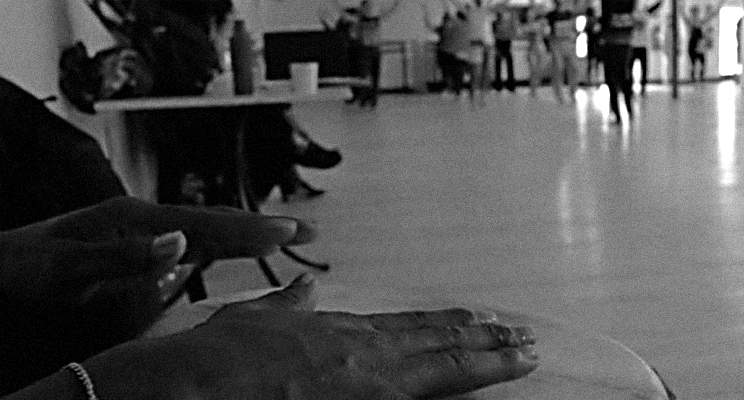 Fotografía en primer plano de unas manos interpretando sobre una conga y de fondo unas bailarinas practicando.