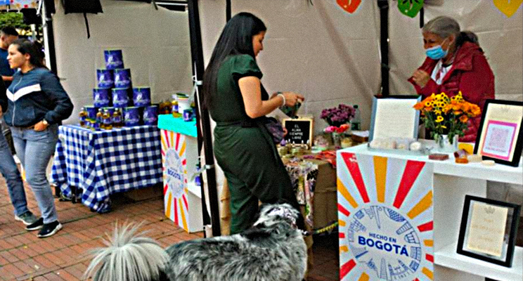 Fotografía de parte de los comercios que hacen parte de la Feria Hecho en Bogotá, en la accion se puede apreciar una mujer joven comprando un producto a una señora de la tercera edad que atiende el estand.