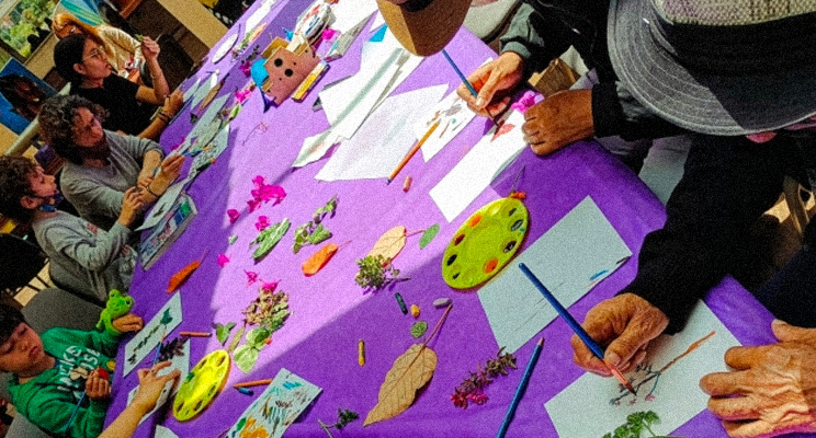 En la foto sobre una mesa de mantel violeta aparecen las manos de un adulto mayor ilustrando una rama de flores rojas y a la izquierda un niño de buzo verde dibujando una planta