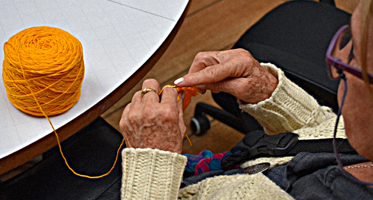 Fotografía de las manos de una señora adulta mayor tejiendo con lana de color amarillo quemado