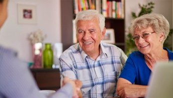 Dos adultos mayores (Mujer y Hombre) sonriendo frente a un asesor que los guia