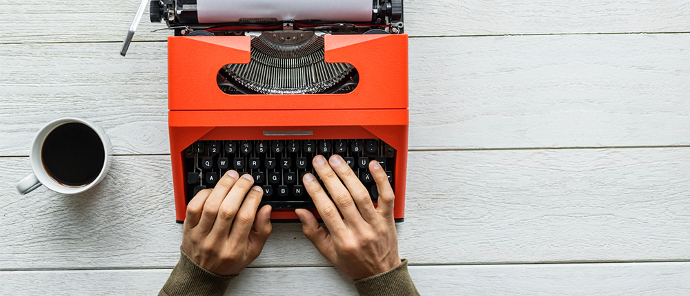 Fotografía en toma cenital de una maquina de escribir de color rojo en la cual hay unas manos adultas dispuestas a escribir y al lado una taza de tinto, todo sobre una mesa de madera blanca.